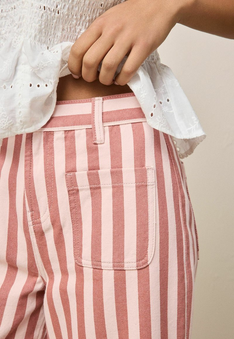 Mano che solleva una camicetta bianca con occhielli, mostrando pantaloni a righe verticali rosa e bianche con dettaglio della tasca posteriore.