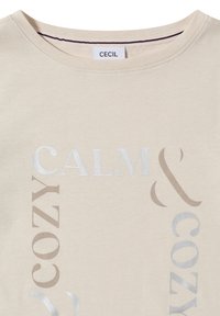 Beżowa bawełniana koszulka z okrągłym dekoltem, z napisami "CALM" i "COZY" w różnych odcieniach srebra i taupe, podkreślająca minimalistyczny design.