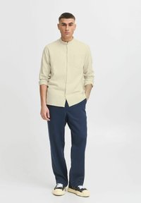 Helles beiges Hemd mit Stehkragen, kombiniert mit locker sitzenden, marineblauen Hosen. Das Outfit wird mit lässigen schwarzen Sneakers vervollständigt.