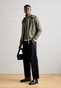 Olivgrüne Jacke mit weißen Kordeln, über einem dunkelgrünen Pullover getragen. Schwarze Hose und Lederstiefeletten. Kleine schwarze Tasche in der Hand.