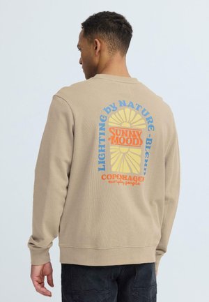Homme portant un sweat-shirt beige avec un graphique solaire coloré "Sunny Mood" et le texte "Lighting by Nature Blend Copenhagen everyday people" au dos.