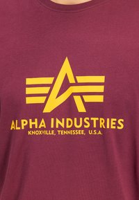 Sárga Alpha Industries logó és szöveg egy bordó pólón, rajta az felirat: „Knoxville, Tennessee, USA.”