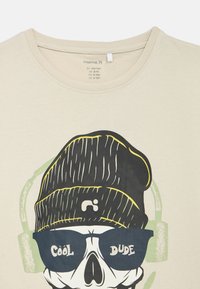 Bēšas krāsas kokvilnas t-krekls ar grafiku, kurā ir galvaskauss ar melnu cepuri un saulesbrillēm, un tekstu "COOL DUDE" treknā baltā krāsā.