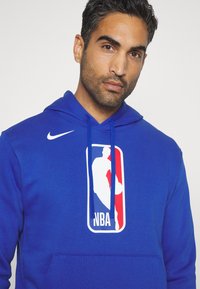 Niebieska bluza z kapturem z frontową kieszenią, na której widnieje duże logo NBA w białym, czerwonym i niebieskim kolorze; zawiera kaptur ze sznurkiem oraz swoosh Nike.