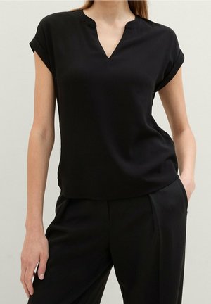 Kobieta w krótkim czarnym bluzce z wycięciem przy dekolcie, w połączeniu z czarnymi spodniami, stoi na jednolitym tle.