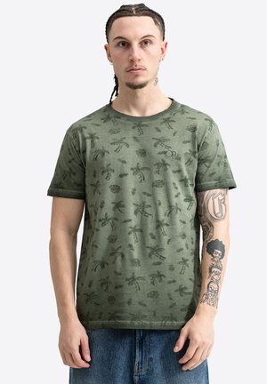 Jeune homme aux cheveux tressés portant un t-shirt vert imprimé de palmiers et un jean, montrant des tatouages sur son avant-bras droit.