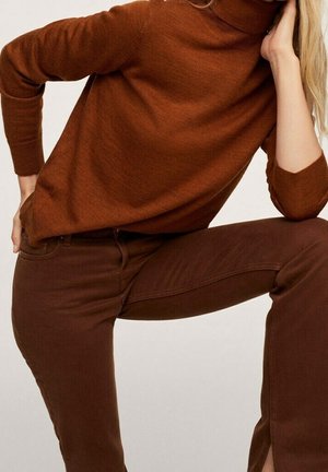 Personne portant un pull marron à manches longues et un pantalon marron assorti, avec un genou relevé et la main reposant sur le visage.