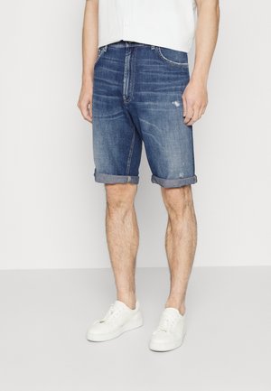 Denim shorts in dark blue, featuring a distressed finish and rolled hems. Paired with white sneakers, on a light background.