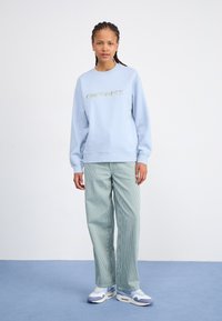 Lys blå hettegenser med "carhartt."-logo i gull, sammen med grønne stripete vide bukser og hvite sneakers med blå detaljer.