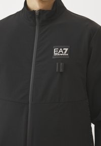 Svart zip-up jacka i mjukt tyg, med en märkespatch på bröstet, hög krage och subtila texturerade detaljer på sidorna.