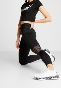 T-shirt nera corta con logo Puma bianco, abbinata a leggings neri con inserti in mesh e stampa sul polpaccio. Sneakers bianche con suola animalier.