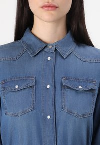 Chemise en denim bleu clair, avec un devant à boutons, un col, deux poches poitrine, et des boutons perlés avec des détails de couture contrastants.
