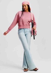 Sweatshirt corto rosa, jeans a zampa d'elefante blu chiaro e sandali neri. Gli accessori includono una borsa a fantasia e un foulard rosa stampato.