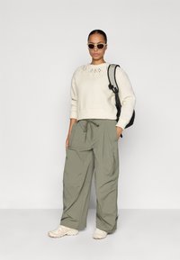 Jersey crema con patrón de diamantes en el escote; pantalones cargo verde oliva con bolsillos; zapatillas blancas; mochila negra; gafas de sol de gran tamaño.