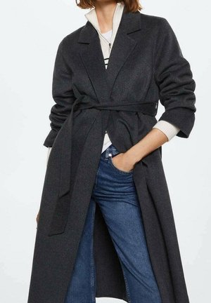 Femme portant un long manteau gris foncé ceinturé par-dessus un haut blanc zippé et un jean bleu, avec une main dans la poche.