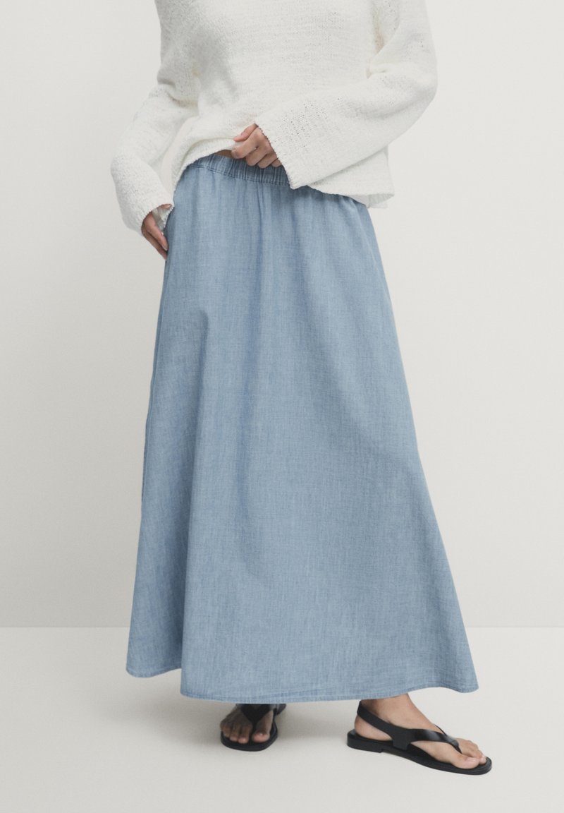 Donna che indossa un maglione bianco a maniche lunghe, una gonna azzurro chiaro fino alla caviglia con vita elastica e sandali neri flat.