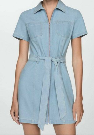 Femme portant une robe en denim bleu clair à manches courtes avec fermeture éclair devant, deux poches poitrine et une ceinture nouée à la taille sur un fond uni.