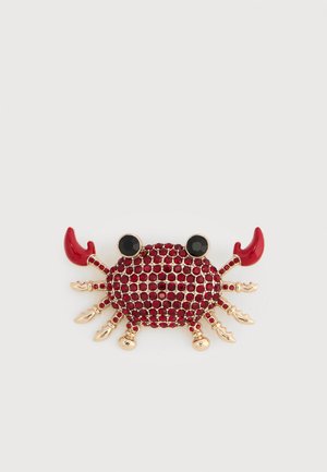 Broche crabe en ton or avec strass rouges, yeux noirs et pinces émaillées rouges sur fond blanc.