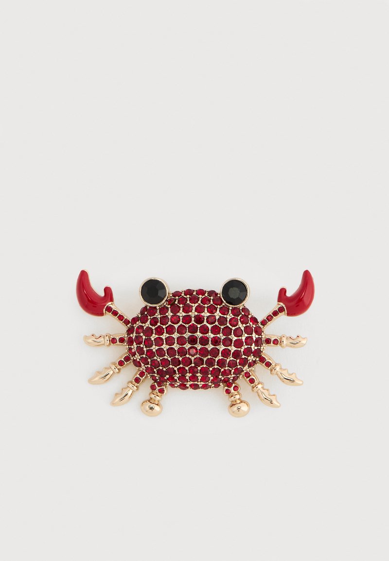 Broche crabe en ton or avec strass rouges, yeux noirs et pinces émaillées rouges sur fond blanc.
