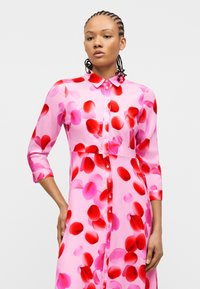 Γυναίκα με πλεξούδες, φορούσα ροζ φόρεμα με κουμπιά και κόκκινα και ροζ μοτίβα από πέταλα, στέκεται μπροστά σε απλό φωτεινό φόντο.