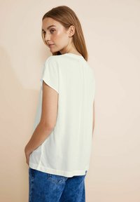 Blusa bianca a maniche corte con vestibilità rilassata, texture morbida e una leggera caduta. La parte posteriore presenta una cucitura centrale e un orlo arrotondato.