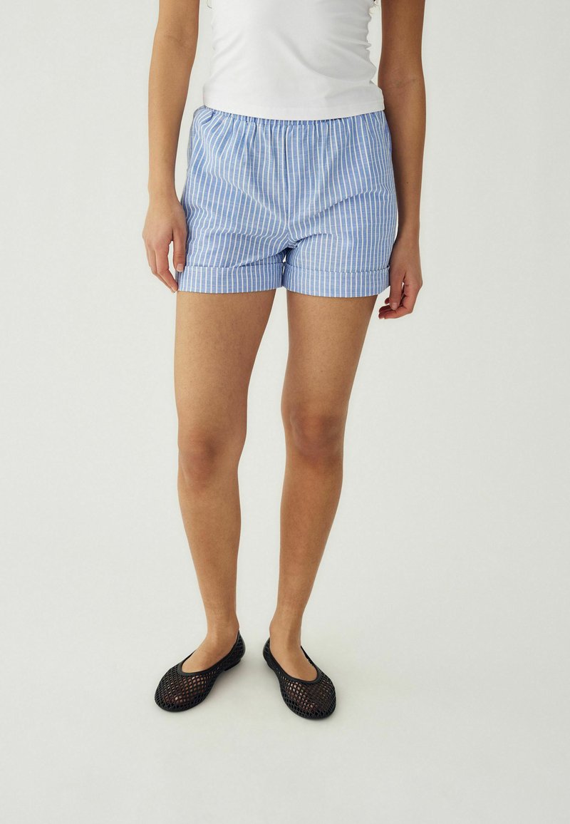 Persona che indossa pantaloncini a righe blu e bianche, top bianco e scarpe slip-on nere in rete, in piedi contro uno sfondo semplice.