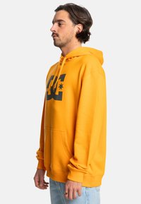 Žltá mikina s kapucňou, veľkým čiernym logom, sťahovacou kapucňou, prednou vreckovou kapsou a rebrovanými manžetami. Hladká textúra látky, ležérny dizajn.