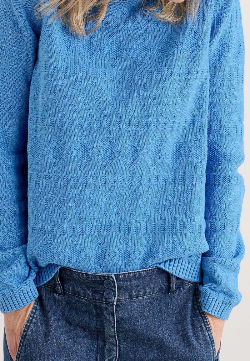 Pull en maille bleu avec motifs géométriques texturés, porté rentré dans un jean bleu foncé taille haute avec fermeture à double bouton.