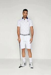 Λευκό πουκάμισο πόλο με διαμαντένιο μοτίβο και ναυτικό γιακά, συνδυασμένο με λευκά σορτς. Τα αξεσουάρ περιλαμβάνουν μια γκρι ζώνη και ένα γκολφ κλαμπ, φορεμένα με αθλητικά παπούτσια.