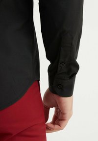 Chemise noire à manches longues avec des poignets boutonnés, texture lisse et ourlet arrondi, vue avec un pantalon rouge. Coutures détaillées sur les manches visibles.