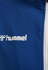 Blått texturerat tyg med ett rutmönster. Har en vit "hummel"-logotyp. Rent design med vita accenter vid kanterna.