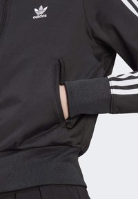 Chaqueta deportiva negra de Adidas con bolsillo con cremallera, puños acanalados y tres franjas blancas a lo largo de las mangas. Logo en el pecho.