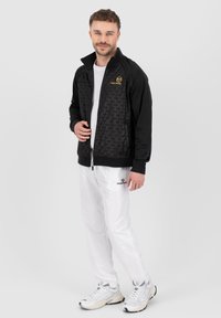 Sort zip-up jakke med et struktureret geometrisk mønster på forsiden, parret med hvide bukser og sølvfarvede sneakers.
