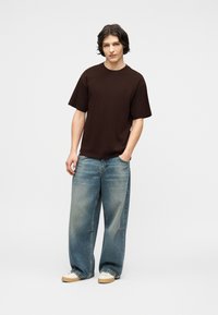 Νεαρός άνδρας με μεσαίου μήκους σκούρα μαλλιά, που φοράει σκούρο καφέ υπερμεγέθη μπλούζα, φαρδιά μπλε τζιν και λευκά αθλητικά, στέκεται με το ένα χέρι στην τσέπη.