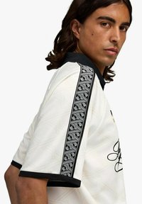 Maglia bianca con motivo a diamante testurizzato, accenti neri e dettagli ricamati. Include una striscia a motivi bianca e nera sulla spalla.