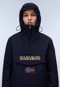 Person trägt eine marineblaue Kapuzenjacke mit einem Reißverschluss am hohen Kragen sowie ein Napapijri-Logo und die norwegische Flagge auf der Fronttasche.
