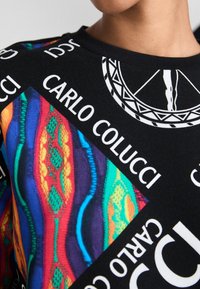 Svart hettegenser med et fargerikt lappeteppe-design med abstrakte mønstre og tekst, inkludert "CARLO COLUCCI" i fete hvite bokstaver.