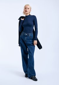 Navyblaues Langarmoberteil kombiniert mit lockeren, dunkelblauen Jeans. Das Model hält eine schwarze Oberbekleidung. Schwarze Schuhe komplettieren das Outfit.