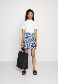 Top branco ajustado com mangas curtas, acima de uma saia com padrão floral azul. Uma bolsa de mão preta texturizada e sandálias planas pretas completam o visual.