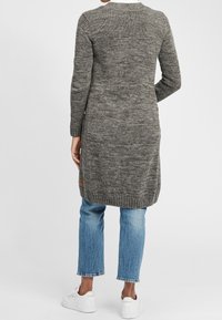 Strickjacke in einem dunklen Grauton, mit langen Ärmeln und einem leicht verlängerten Rücken. Auffälliges Etikett-Detail am Saum; kombiniert mit blauen Jeans.