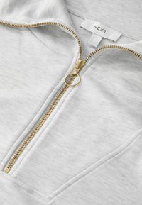 Sweat à capuche gris clair zippé avec une fermeture éclair dorée et une languette, présentant un tissu texturé et une petite étiquette portant l'inscription "NEXT".