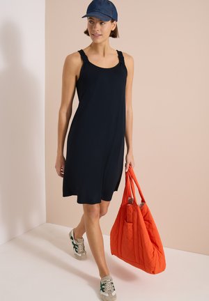 Femme marchant à l’intérieur portant une robe bleu marine, une casquette bleue, des baskets blanches, et tenant un grand sac cabas matelassé orange vif.