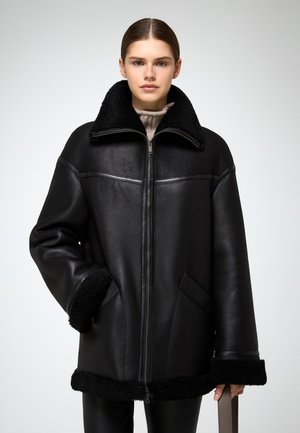 Giacca in pelle nera con collo in pelliccia, cerniera frontale e due tasche laterali. Presenta una consistenza morbida e maniche ampie, abbinata a pantaloni scuri.