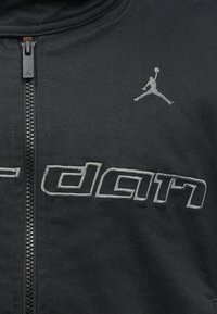 Jaqueta preta com fecho éclair e texto e logotipo "Jordan" bordados. Apresenta uma textura de tecido suave e um design ajustado.