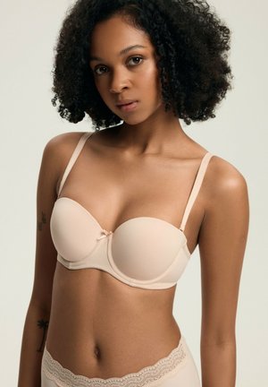 Béžová podprsenka bez ramínek s hladkým materiálom, tvarovanými košíčkami a centrálnou mašľou. Nastaviteľné ramienka a čipkované akcenty pozdĺž dolného okraja.