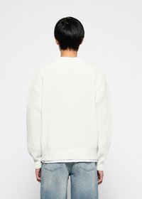 Person mit kurzen schwarzen Haaren, die einen weißen Strickpullover und ausgebleichte blaue Jeans trägt, steht vor einem schlichten weißen Hintergrund.