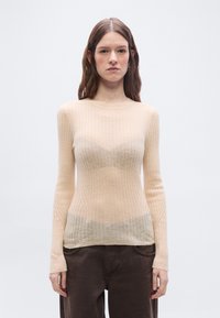 Jeune femme aux cheveux bruns raides portant un pull beige transparent à manches longues côtelé et un pantalon marron foncé, debout devant un fond blanc.