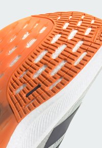 Suela de zapato con una capa de goma naranja con patrones de agarre texturizados, una entresuela blanca y un acento lateral gris.