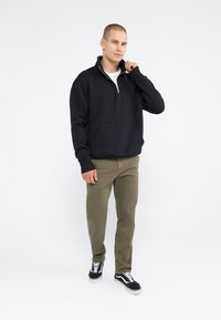 Sweat à zip noir avec une poche kangourou, associé à un pantalon vert olive et des chaussures noires. Texture lisse et coupe décontractée.