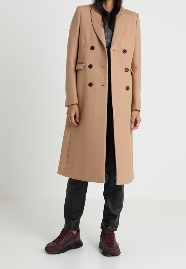 Manteau long beige à double boutonnage avec des revers crantés et des boutons noirs, associé à un pantalon en cuir noir et des sneakers bordeaux.